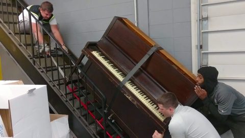 moving crew tilting an upright piano up an industrial stairwell with a strap and piano dolly, face visible