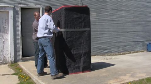 Two-man crew wrapping a moving blanket around an appliance loaded on a red appliance dolly outdoors, face visible