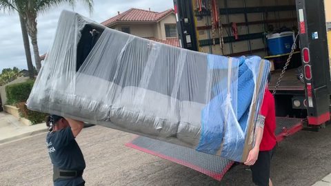 Two-man crew loading a blanket-and-plastic-wrapped sofa onto the liftgate of a moving truck, face visible