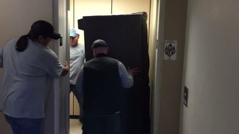 Moving crew guides a wrapped mattress into an apartment building elevator, face visible